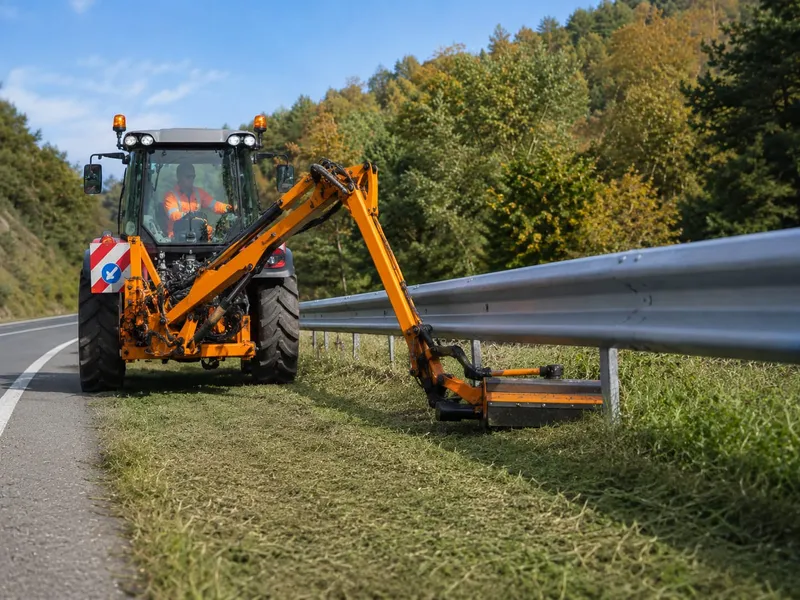 Tracteur avec bras de fauchage entretenant la végétation sous une glissière de sécurité
